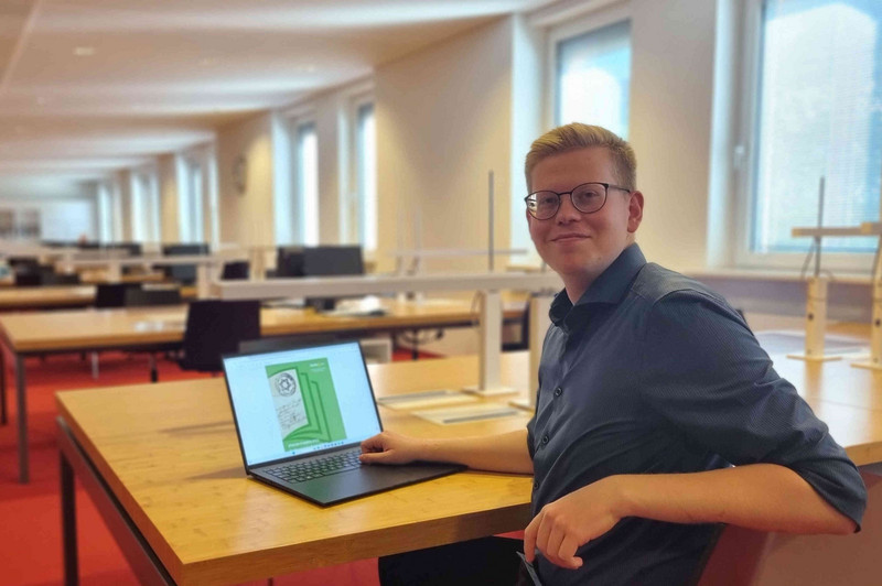 Projektmitarbeiter Daniel Pöhl sitzt an einem Tisch im Lesesaal vor einem Notebook. Auf dem Bildschirm ist das Deckblatt eines Quellennah-Heftes zu sehen. 