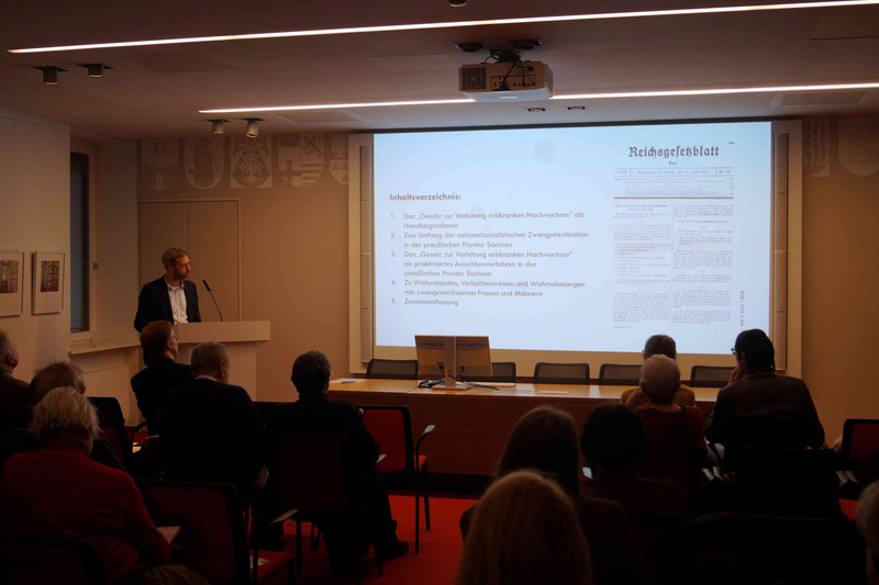 Blick in den Vortragssaal: PD Dr. phil. habil. Alexander Bastian steht während des Vortrags am Rednerpult und verweist auf seine Power-Point-Präsentation. Davor sind die ersten drei Reihen mit Zuhörern zu sehen. 