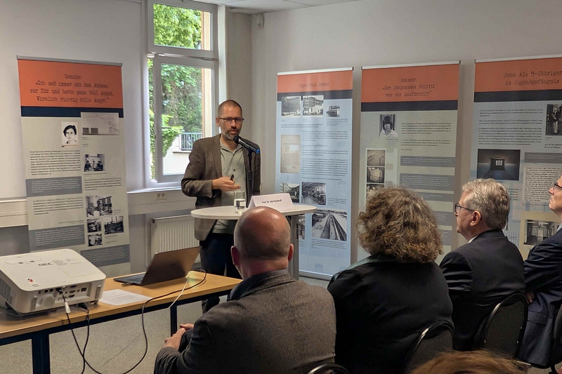 Mann vor Rednerpult mit Publikum im Hintergrund Ausstellungsrollups